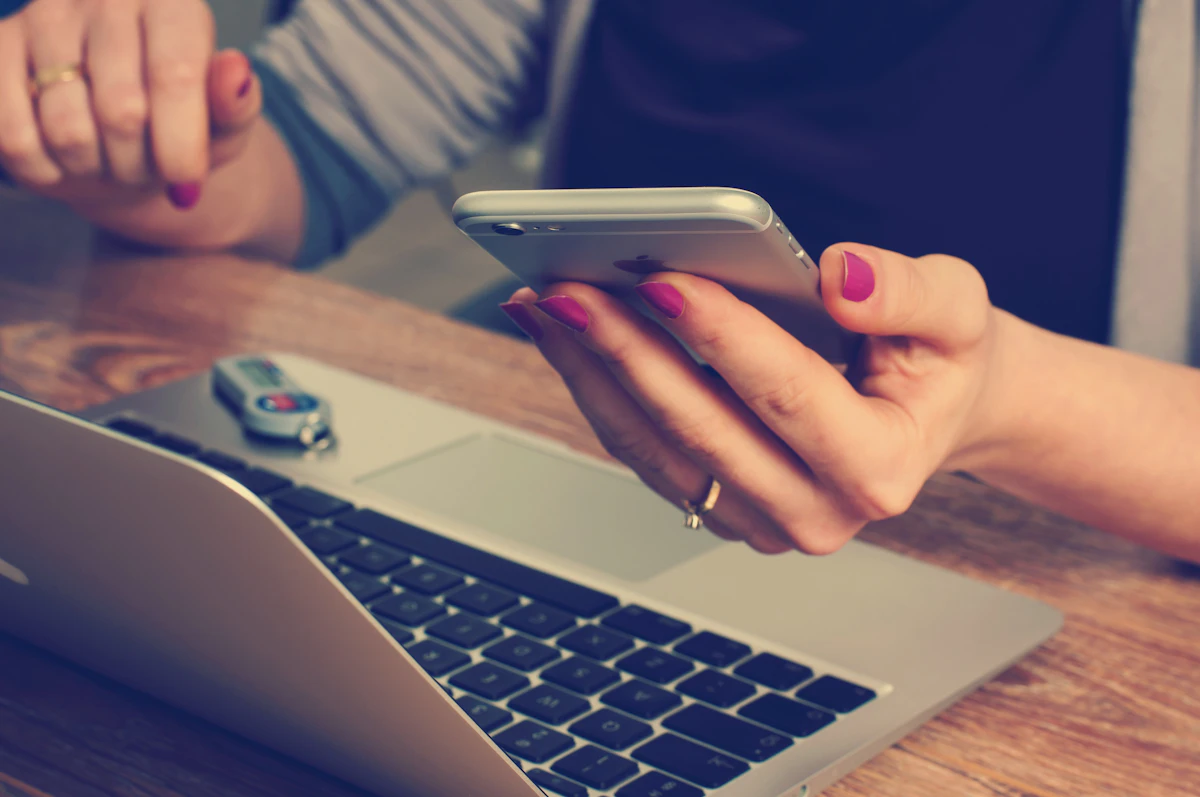 Hands with painted nails holding a smartphone above an open laptop on a wooden desk