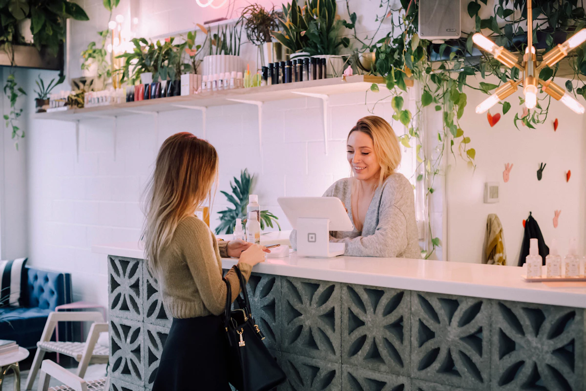 Smiling receptionist at a plant-filled salon front desk with a tablet, serving a customer