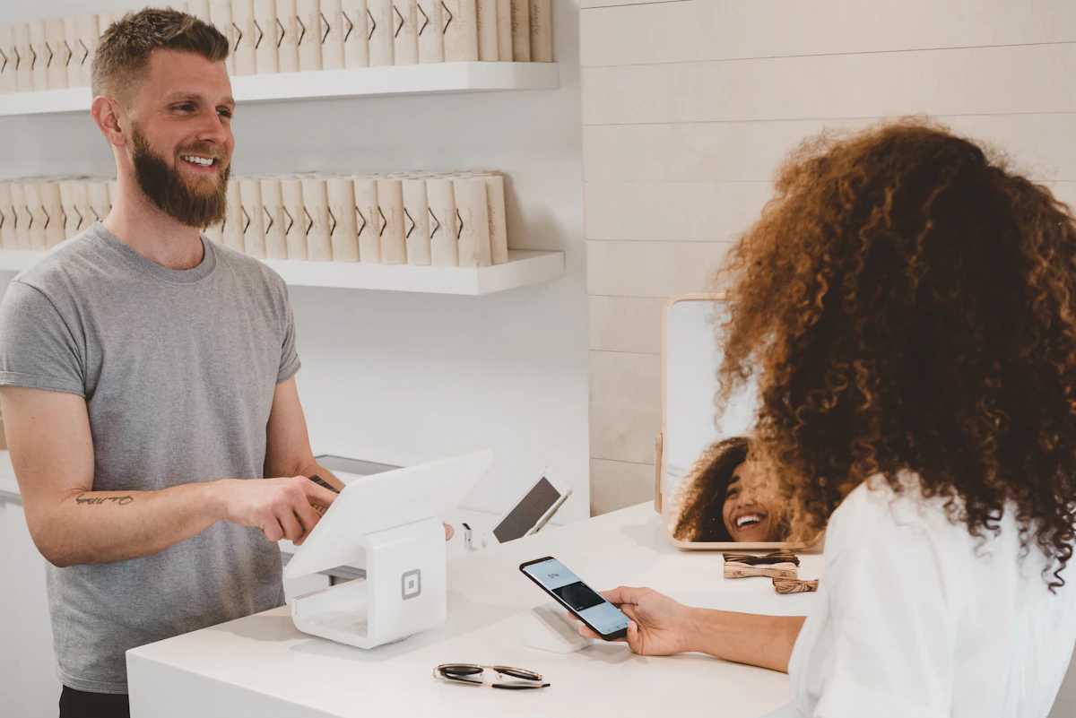 Smiling shop assistant at a white checkout counter with a card terminal as a customer pays with her phone