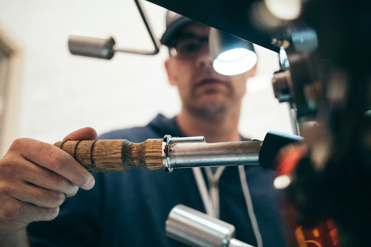 Man in a cap and glasses working a machine by a wooden-handled lever under a small lamp